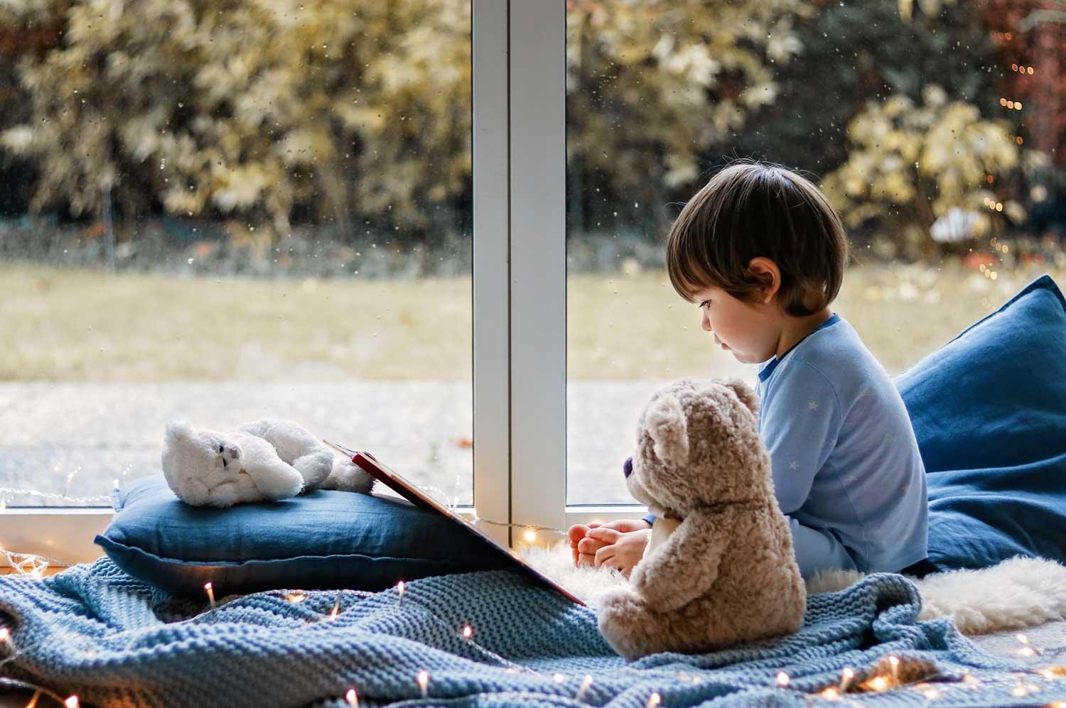cute-little-boy-reading-book-with-his-teddy-bear-toy-sitting-cozy-on-pillows-and-knitted-blanket-near_t20_4bQ14R